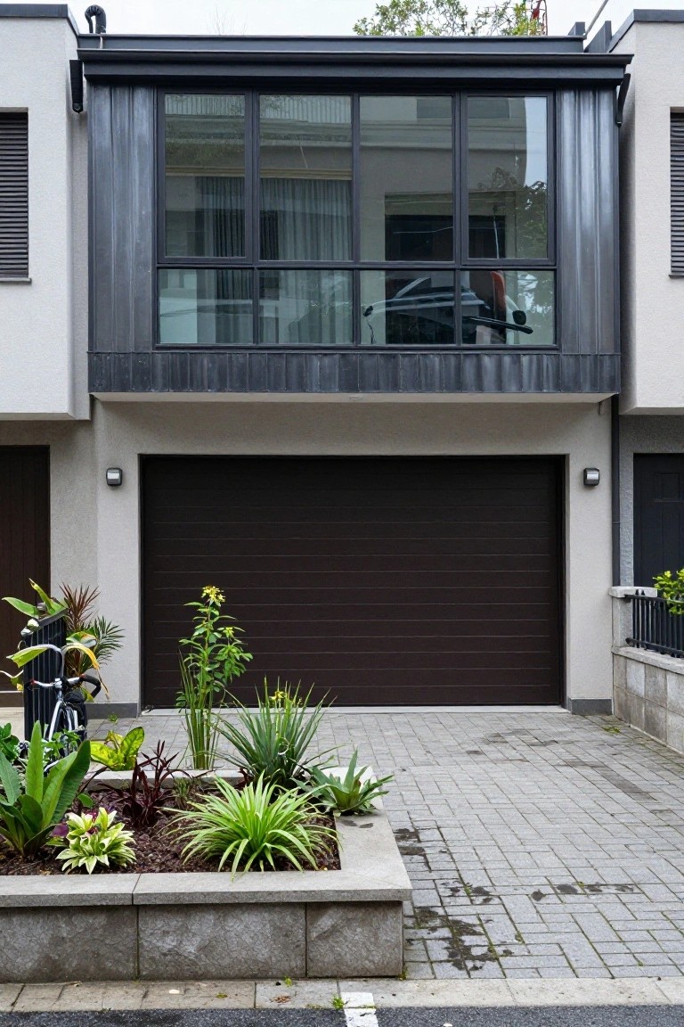 Modern two-story house exterior with light walls, dark brown sectional garage door, dark metal-framed upper balcony windows, raised stone planter beds with tropical plants, and paver driveway.
