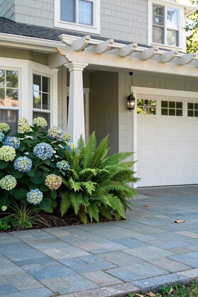 Gray shingled house with white pergola over the garage door, blue hydrangea bushes and ferns planted in front, and a gray paver driveway leading up.