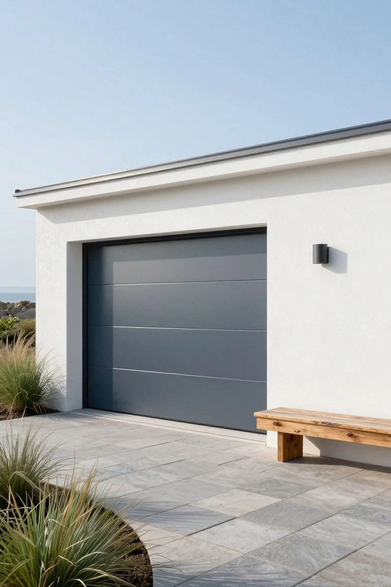 Modern garage facade with dark gray sectional door, white stucco walls, wooden bench, and paver driveway