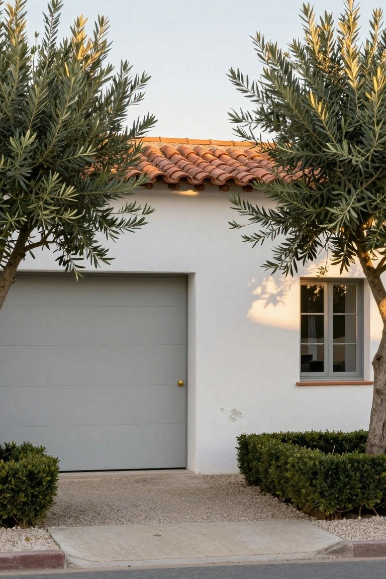White stucco house exterior with large gray garage door, red tile roof, two olive trees flanking the garage, boxwood hedges, and concrete pathway leading to a gray-paver driveway.