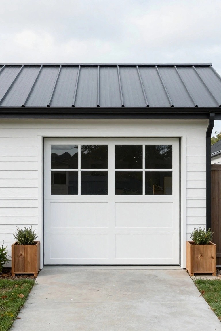 White double garage door with grid windows on white board-and-batten house under dark metal roof, flanked by two tall wooden planters with greenery on concrete driveway.