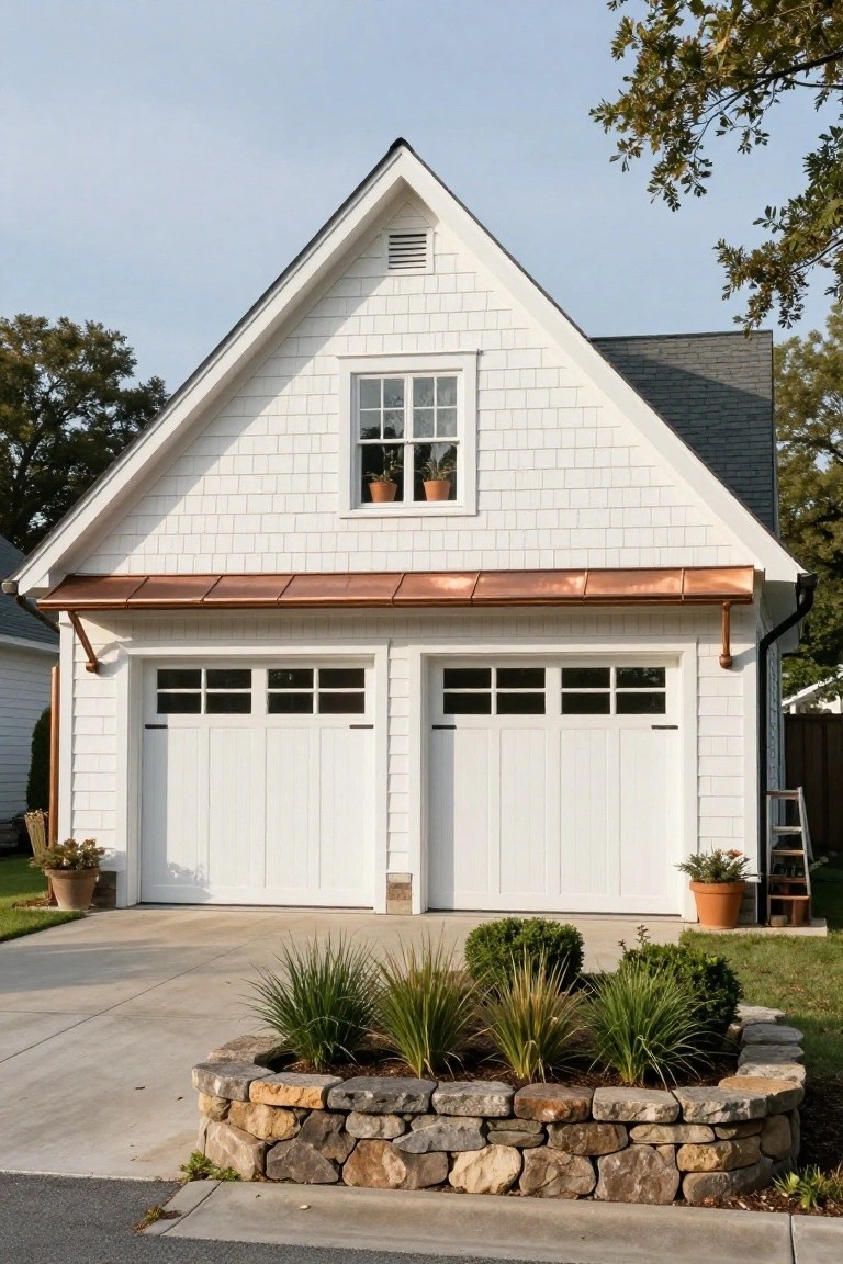 A white detached garage with a steep gable roof, two side-by-side garage doors, a small upper window, and a copper accent above the doors, with a low stone-edged garden bed in front.