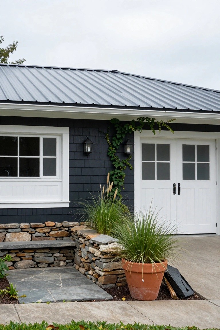 A detached garage with dark shingle siding, white double doors, a white window, and a stone foundation, with plants and a terracotta pot in front.