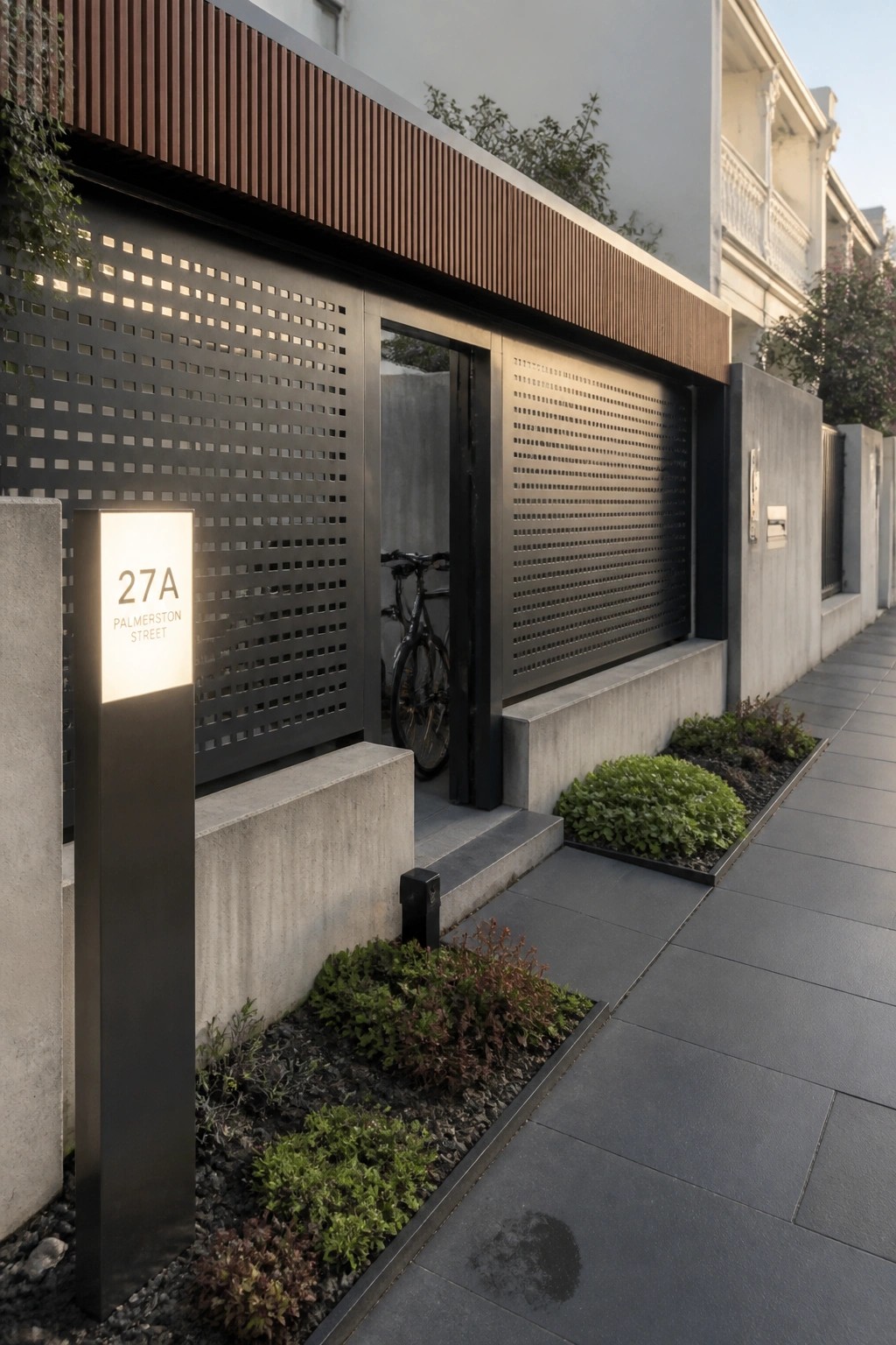 A modern house front with a dark perforated metal sliding gate, concrete landscaping beds, a lit address sign, and a bicycle visible inside the entry.