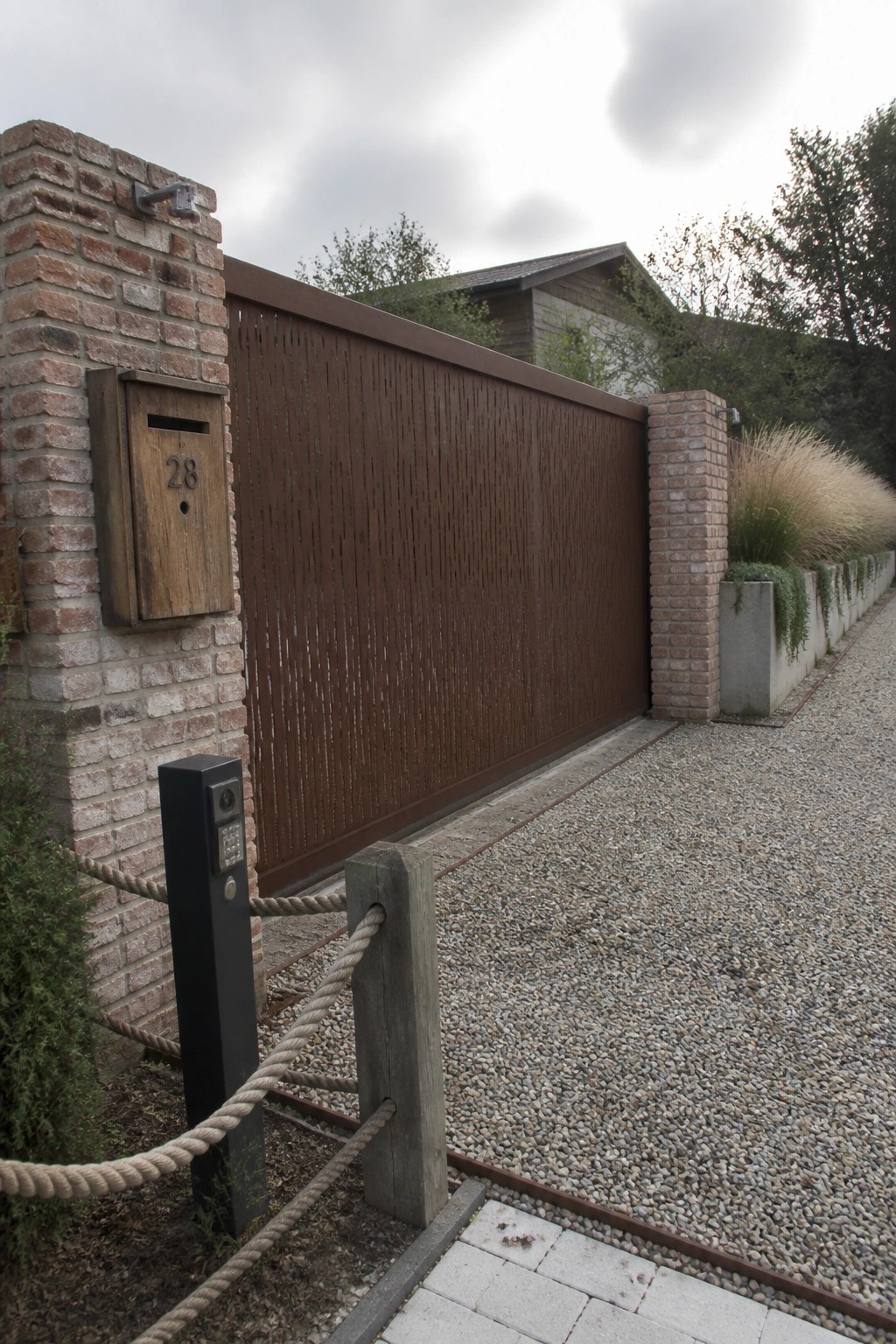 A brown vertical slat sliding gate between brick pillars on a gravel driveway, with a wooden mailbox mounted on the left pillar.
