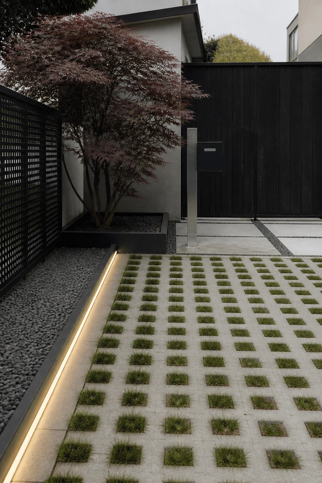 A driveway paved with light concrete grid pavers containing small squares of grass, next to a gravel bed with a glowing linear light and a dark gate in the background.