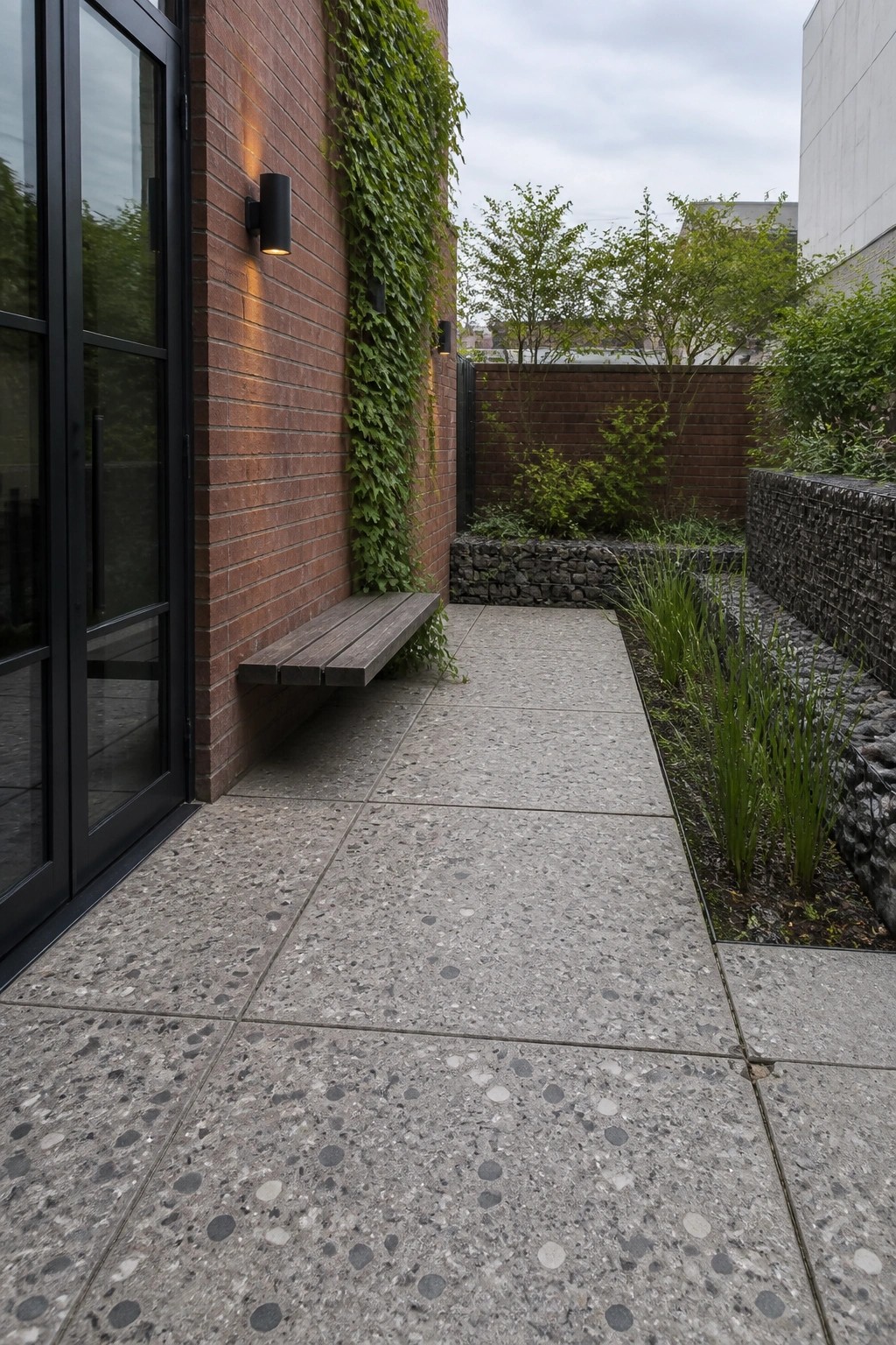 A walkway of large speckled concrete pavers runs between a red brick wall with a wooden bench and a planted bed bordered by gabion walls.