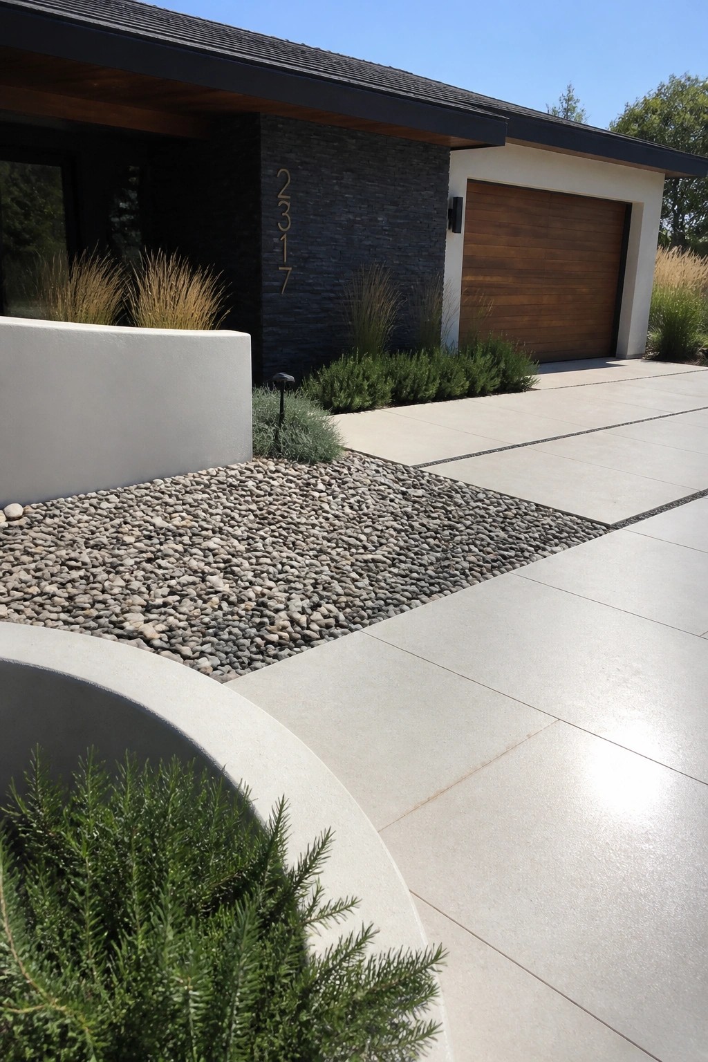 Modern house exterior with a concrete driveway bordered by a wide gravel bed, low shrubs, and ornamental grasses.