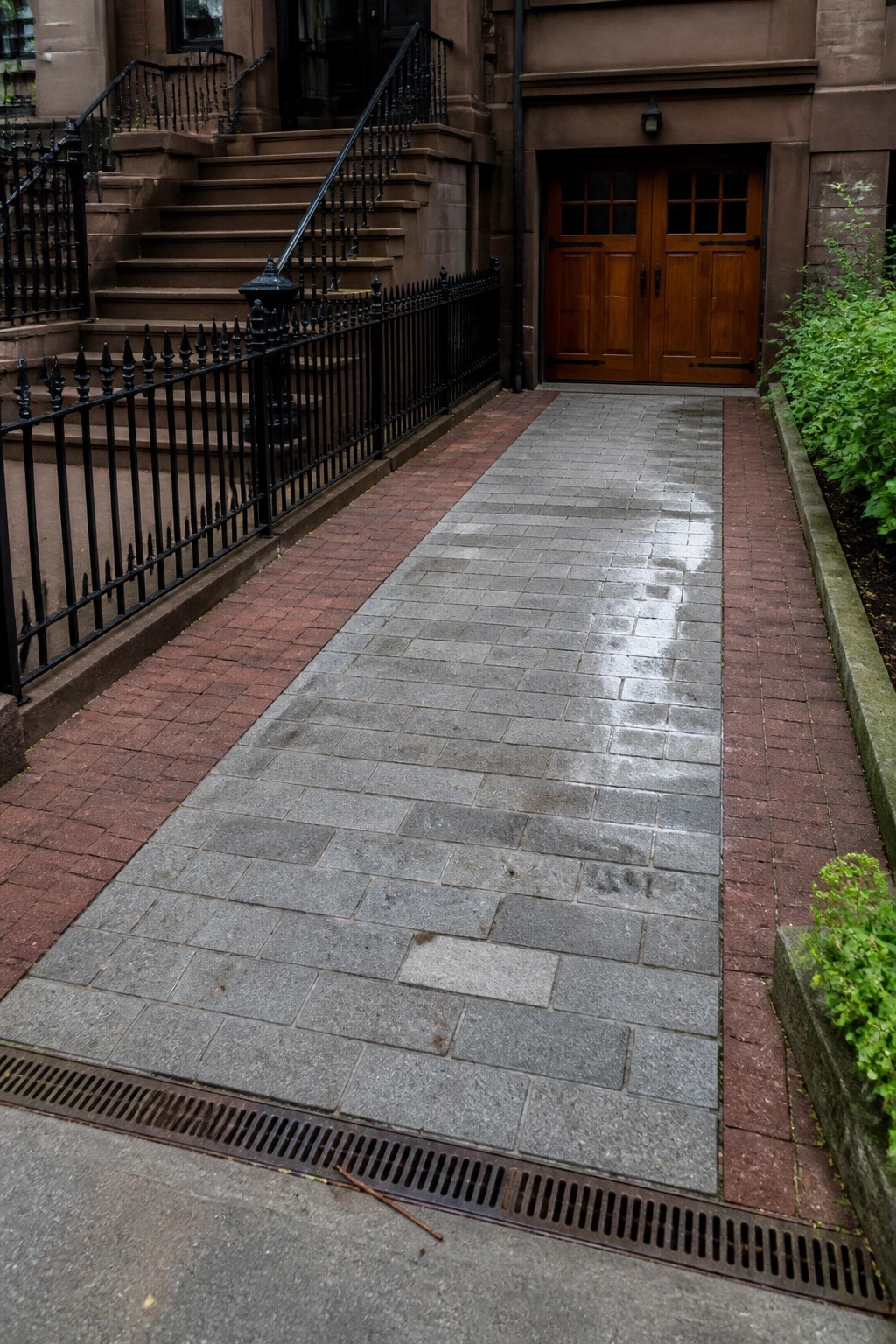 A wet gray stone path bordered by red brick leads to a set of wooden double doors on a brown stone building, with a black metal fence on the left side.