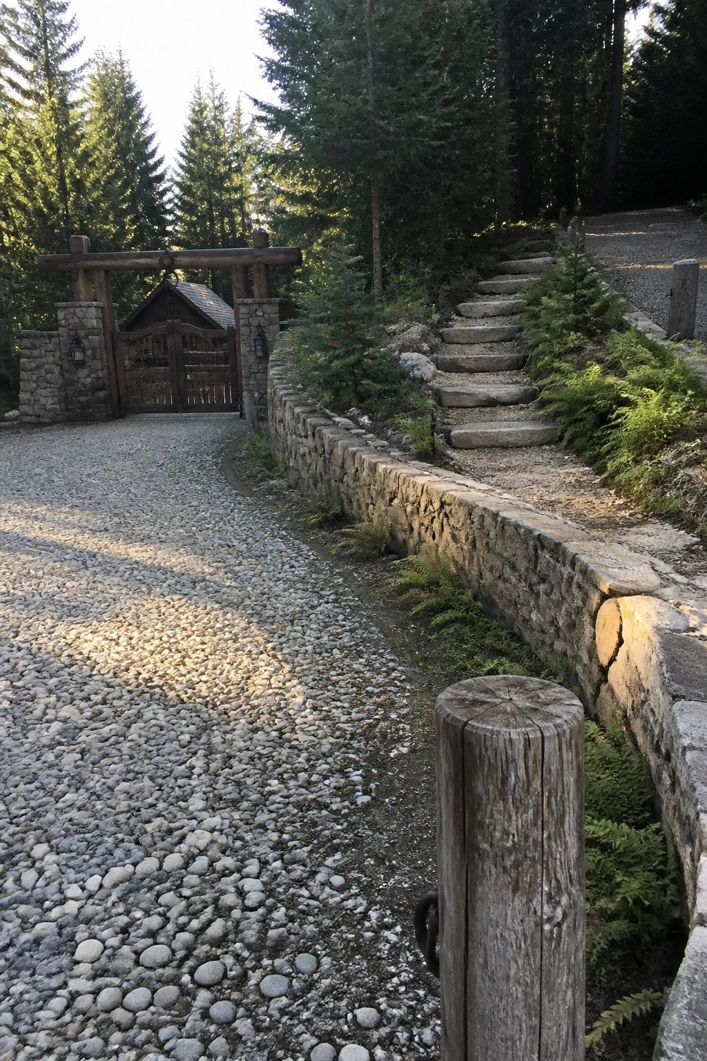 A gravel driveway curves toward a wooden gate between stone pillars, bordered on the right by a low stone wall and stone steps leading uphill through trees.