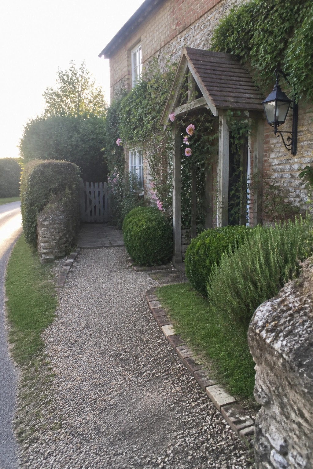 A gravel path bordered with bricks leads past shrubs and a wooden porch on a stone cottage.