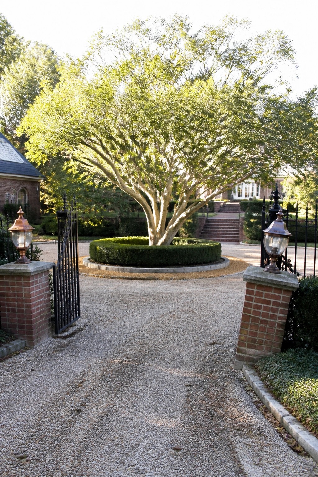 Open wrought-iron gate with lanterns framing gravel drive to central tree and hedge
