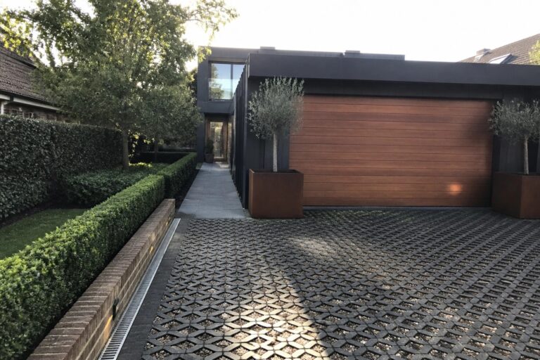 14 Textured Parking Tiles Ideas That Add Depth and Grip Modern house exterior showing a driveway paved with black hexagonal grid tiles filled with gravel, bordered by large rusted metal planters holding small trees.