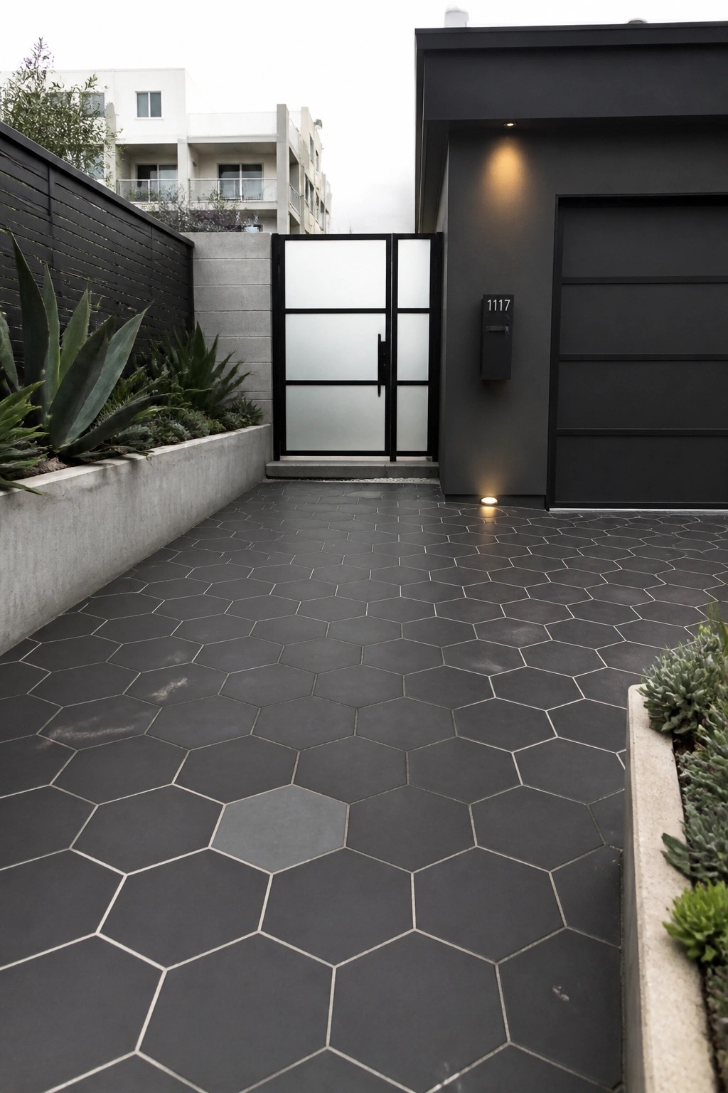Dark gray hexagonal tiles cover a narrow driveway leading to a black metal gate and modern house with a garage door.
