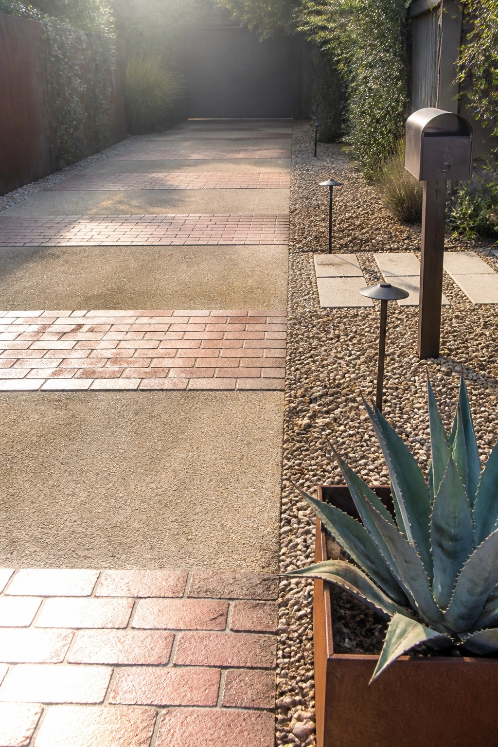 A driveway composed of alternating sections of red brick pavers and gray concrete, bordered by gravel with low landscape lights and an agave plant in a metal planter.
