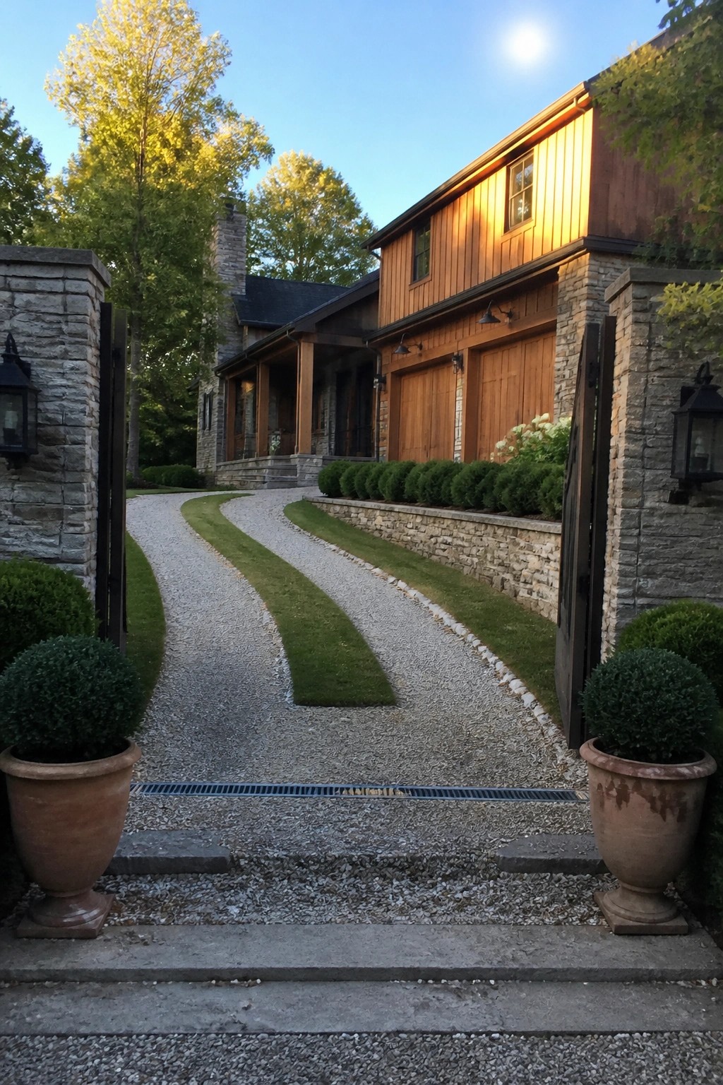 A gravel driveway with two central grass strips curves toward a house with wood siding and stone details, framed by stone gate posts and large terracotta pots.
