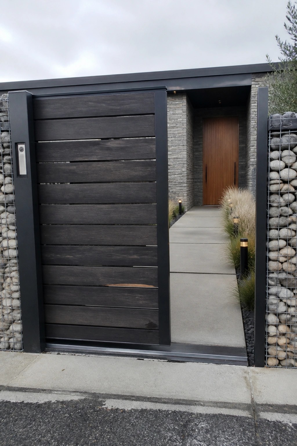 A dark wooden gate with horizontal slats stands open next to gabion stone walls, with a concrete path leading to a wooden door.