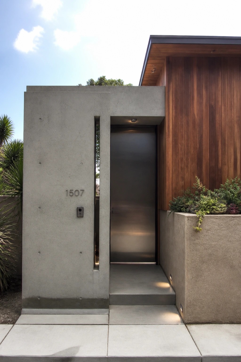 A metallic door recessed into a concrete wall beside vertical wood siding, with a small planter of succulents to the right.