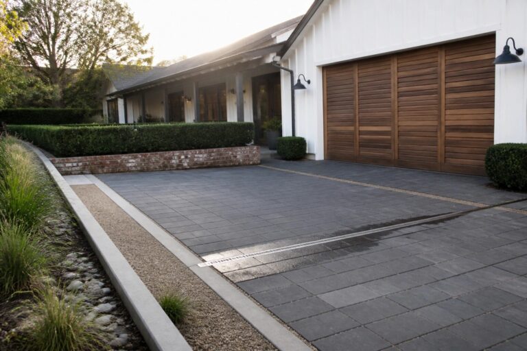 Modern white house with wooden garage door, gray paver driveway, and garden landscaping