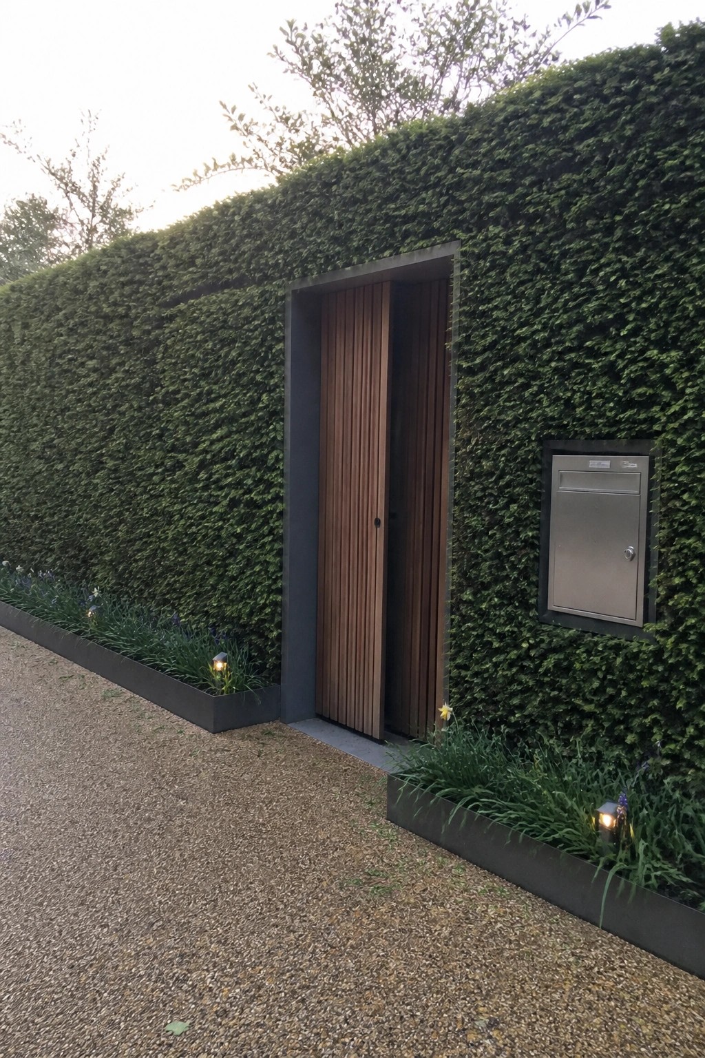 A wooden slatted door set into a tall green hedge wall beside a gravel path, with a metal mailbox mounted to the right and low planters along the base.