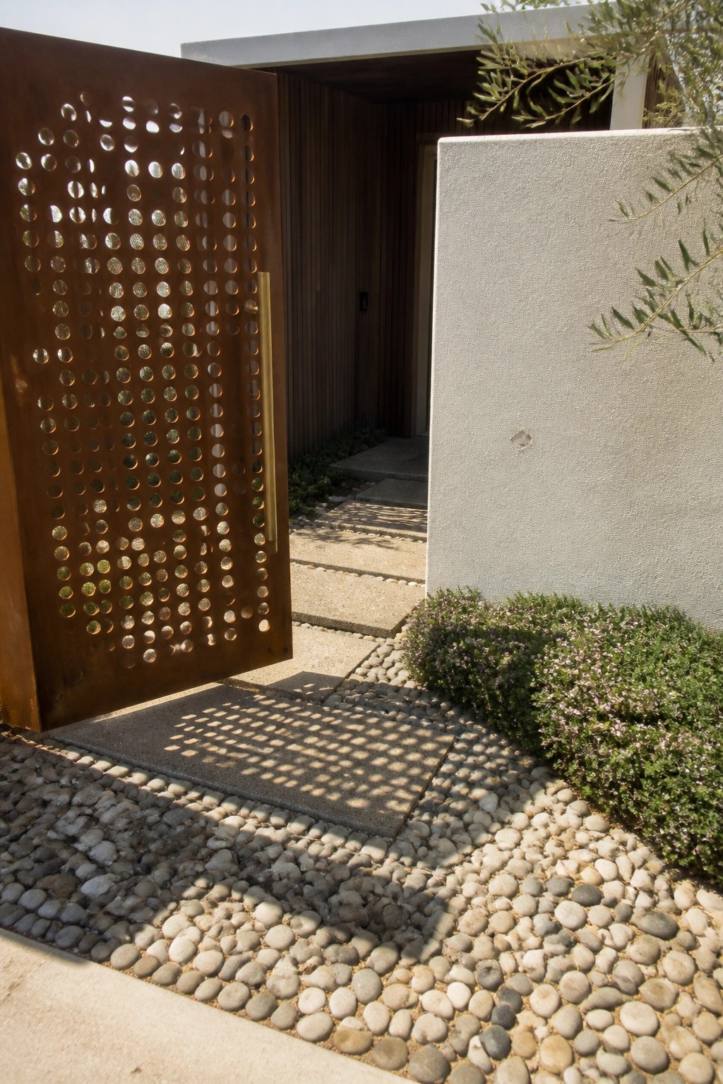 An open rusted metal gate with circular cutouts stands beside a concrete wall next to a pebble path.