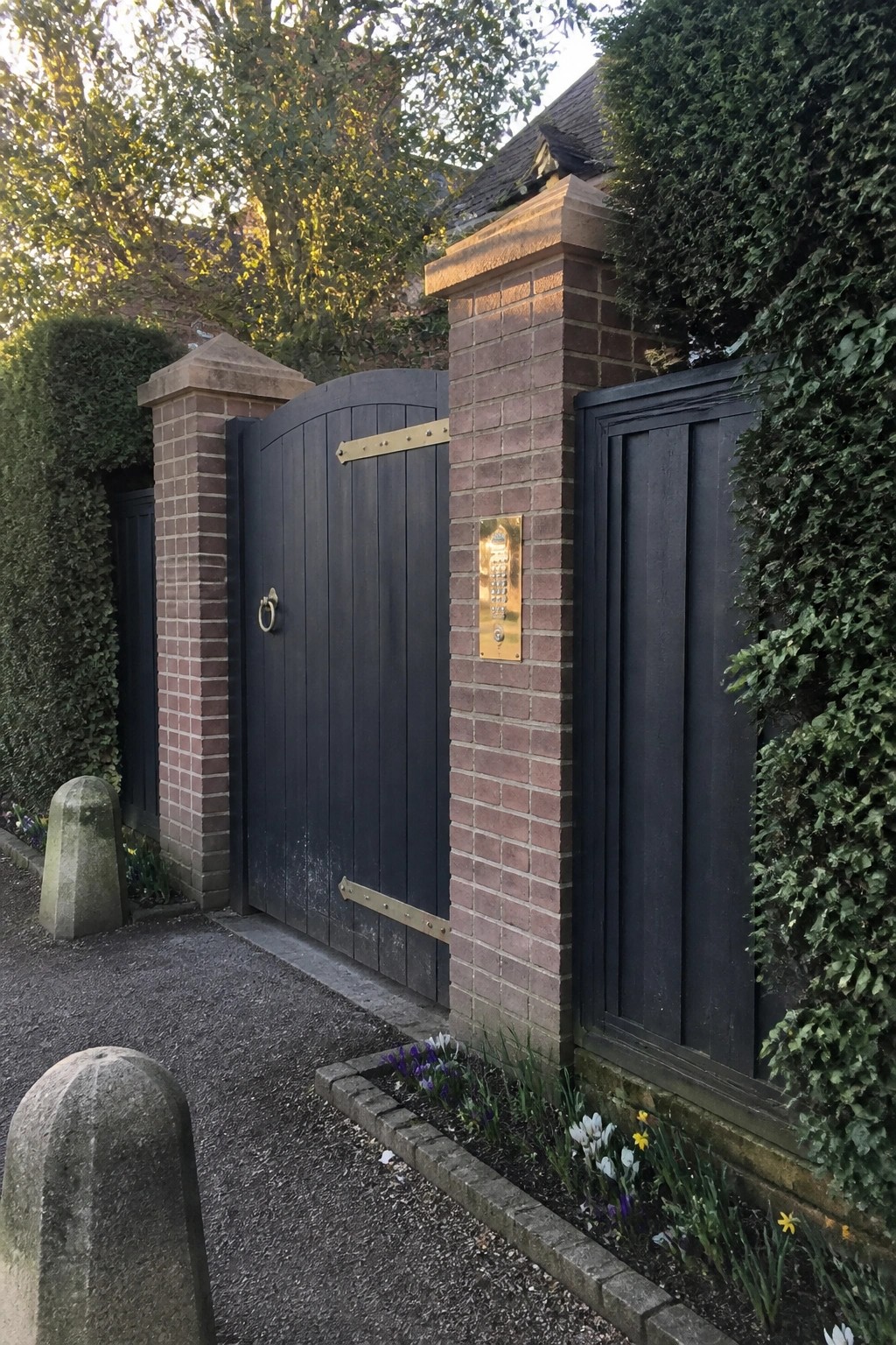 Black wooden gate with brass hinges between brick pillars beside gravel path and flowers
