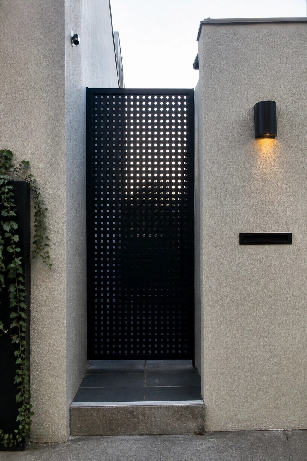 A black perforated metal gate stands between two concrete walls with steps leading up to it, a security camera on the left wall, and a lit wall sconce on the right.