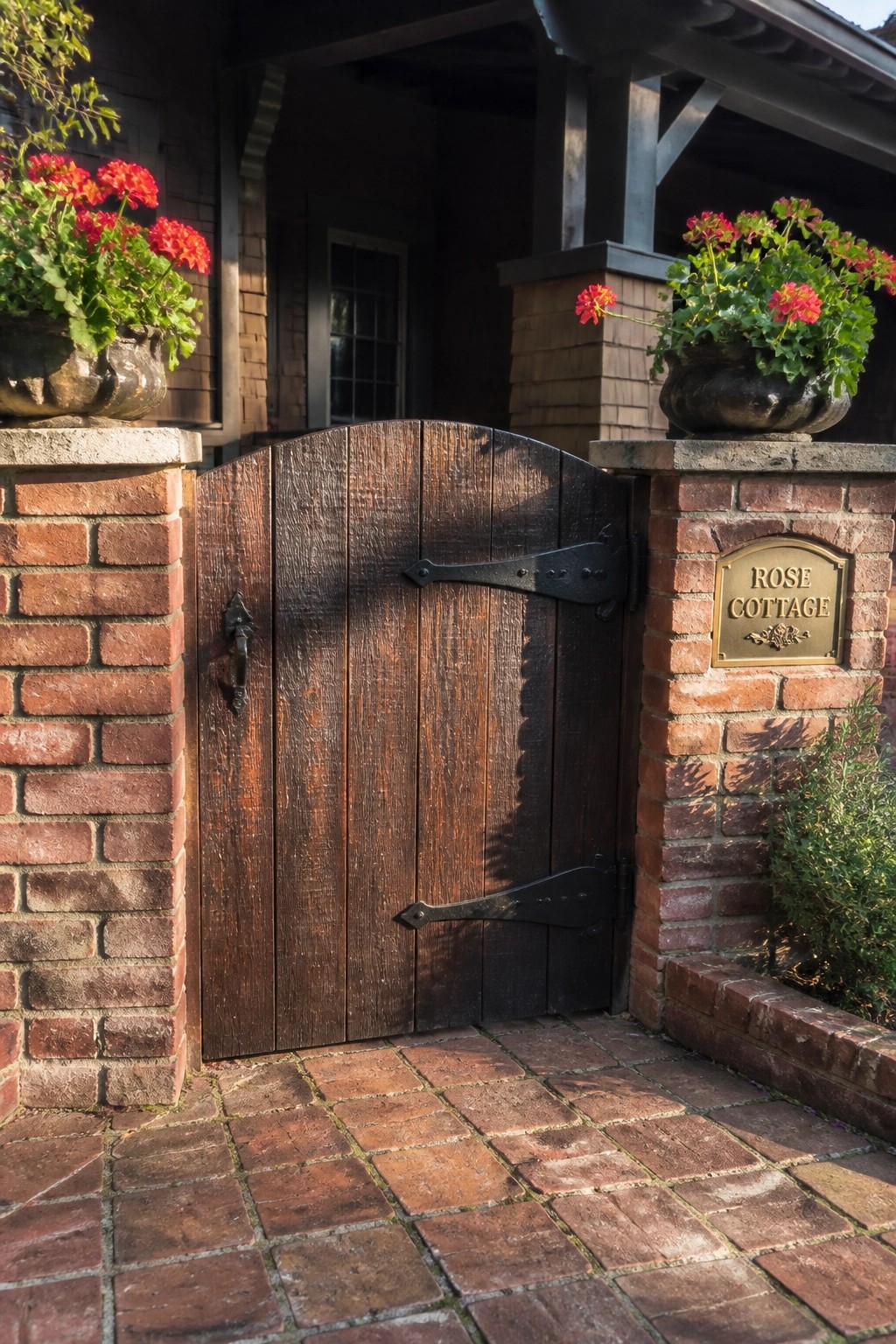 A dark wooden gate with black metal hinges is set between two brick pillars topped with potted red flowers, and a brass plaque on the right pillar reads Rose Cottage.