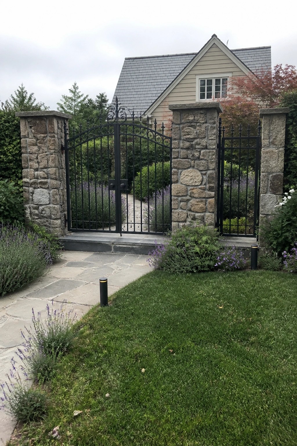 A black wrought iron gate is centered between two tall stone pillars at the front of a house with a stone path and lawn visible beyond.
