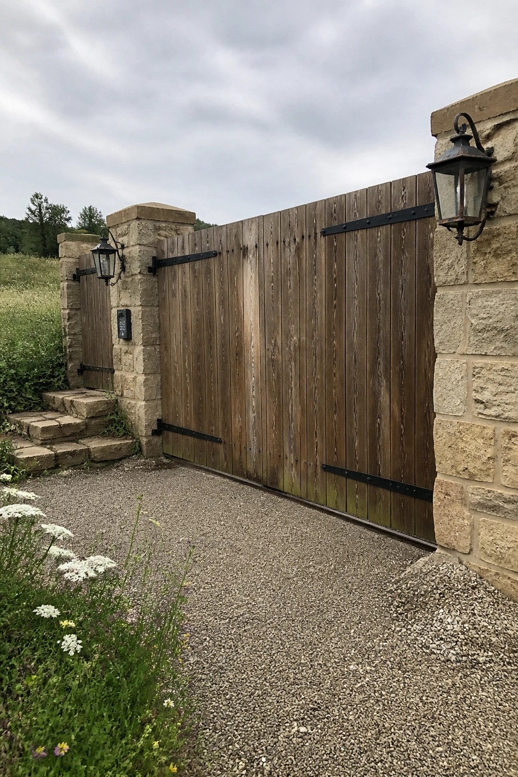 A wooden gate with black metal straps between two stone pillars on a gravel driveway, with lanterns and a keypad visible.