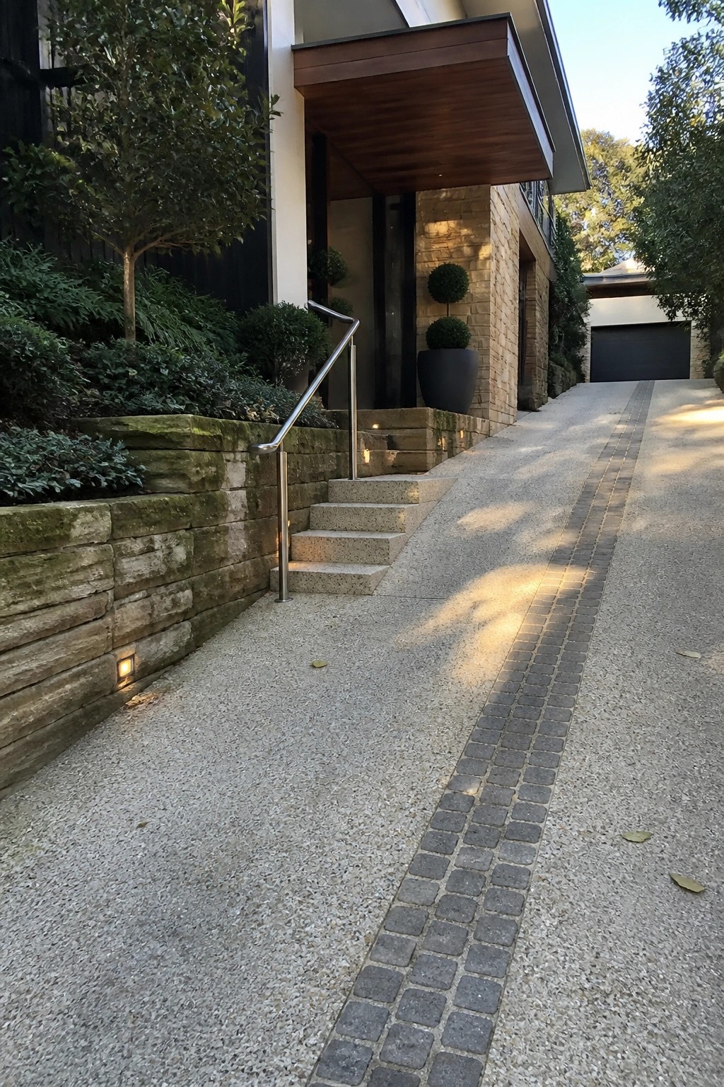 A modern home entrance showing a concrete driveway with a central strip of dark gray cobblestone pavers, stone steps with metal railings, and low landscaping along a retaining wall.