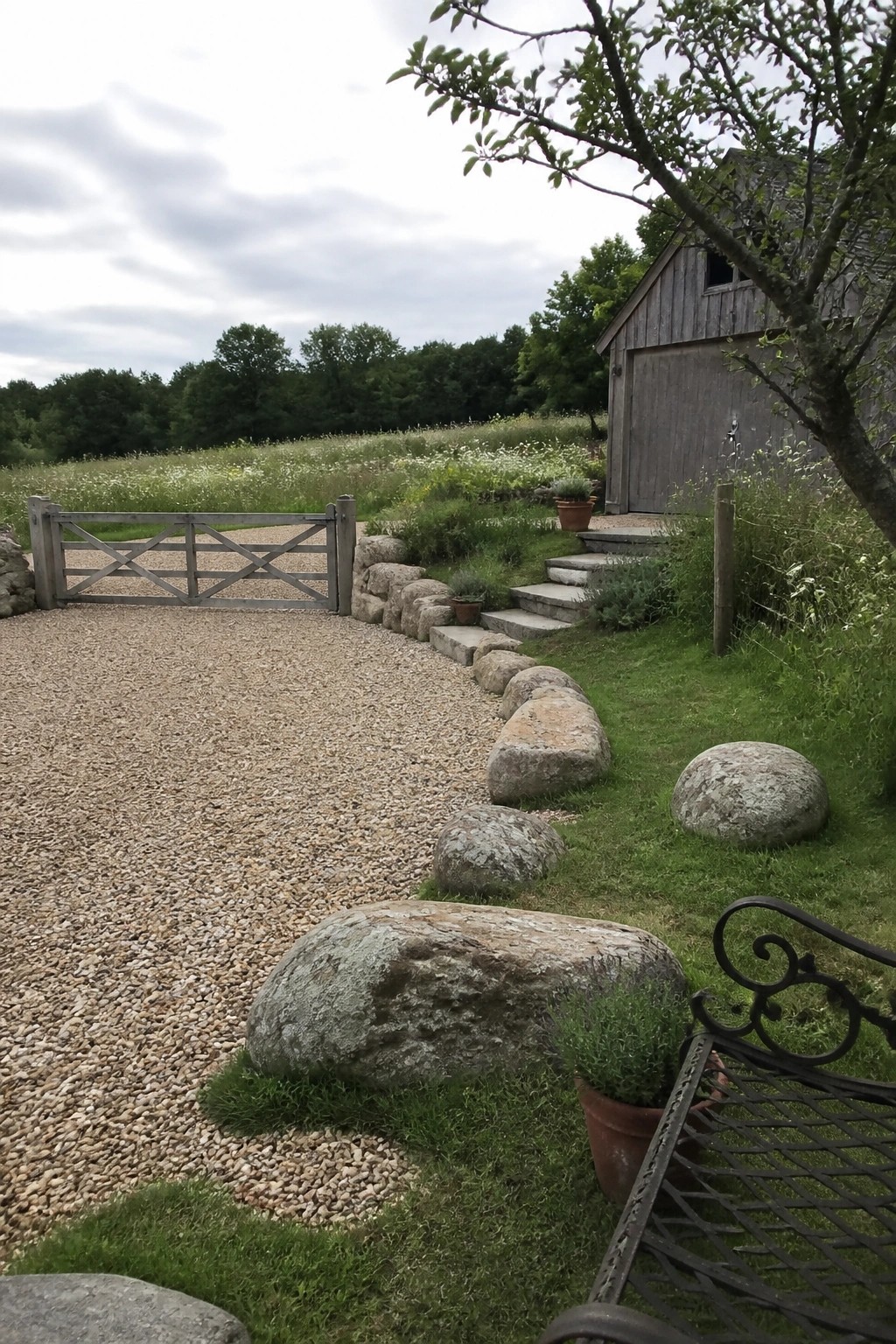 A gravel driveway curves past large stones that form the edging, with a wooden gate on the left and a gray wooden barn on the right.