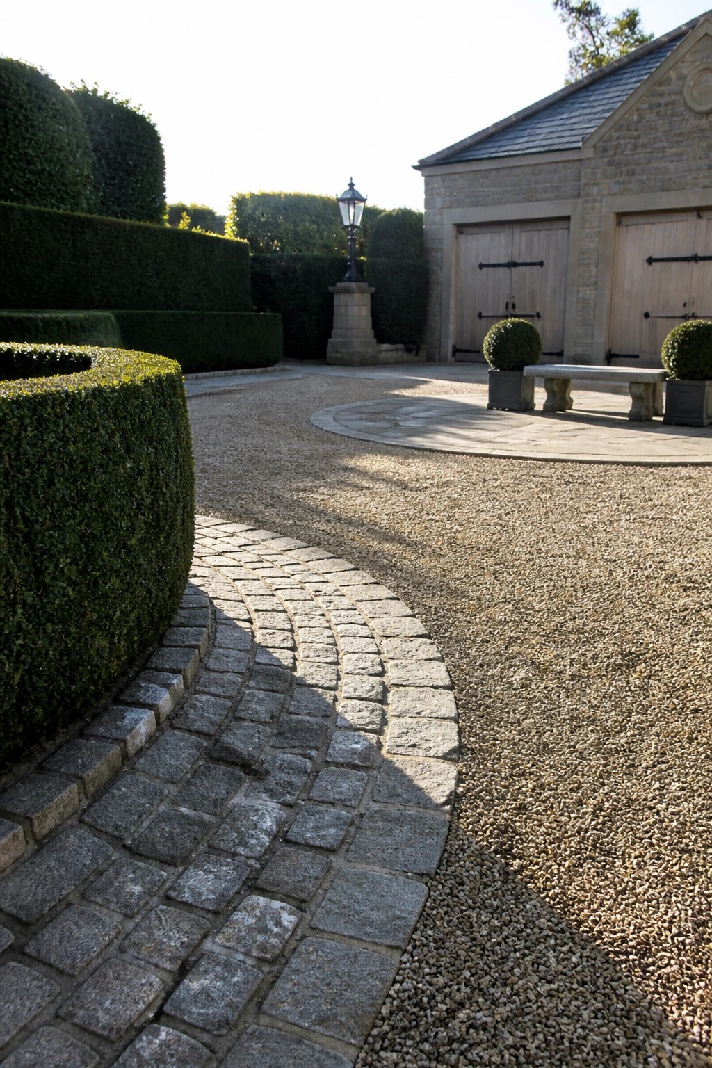 Curved cobblestone path borders gravel courtyard with hedges, stone bench, and garage doors