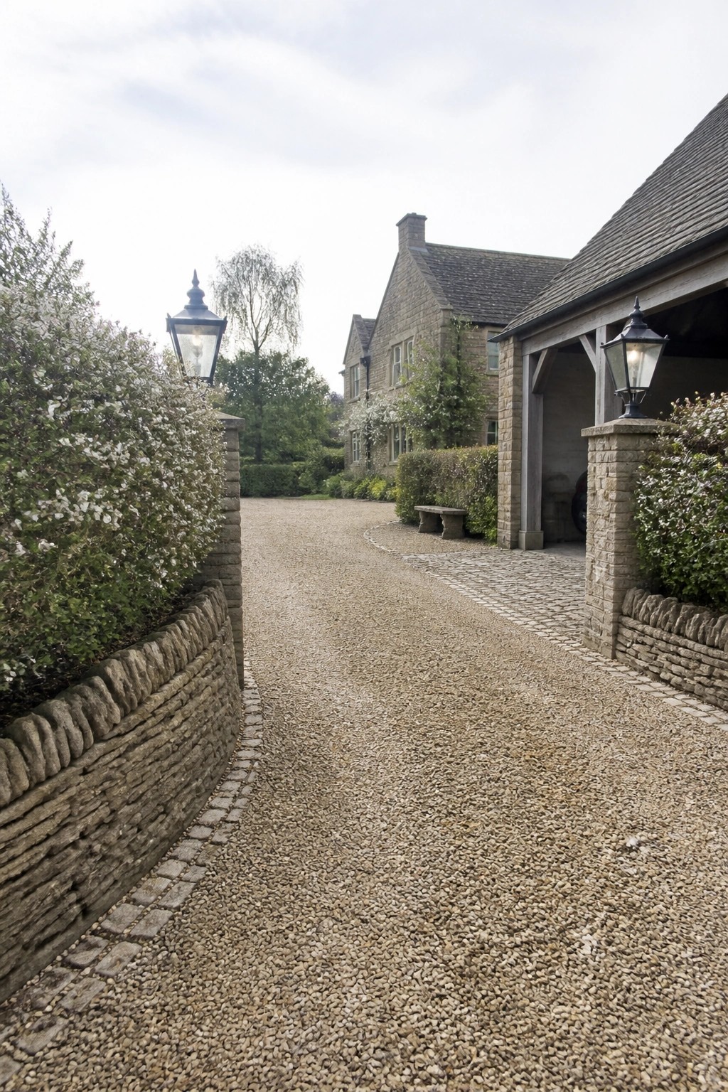 Gravel driveway flanked by stone walls with lanterns leading to house