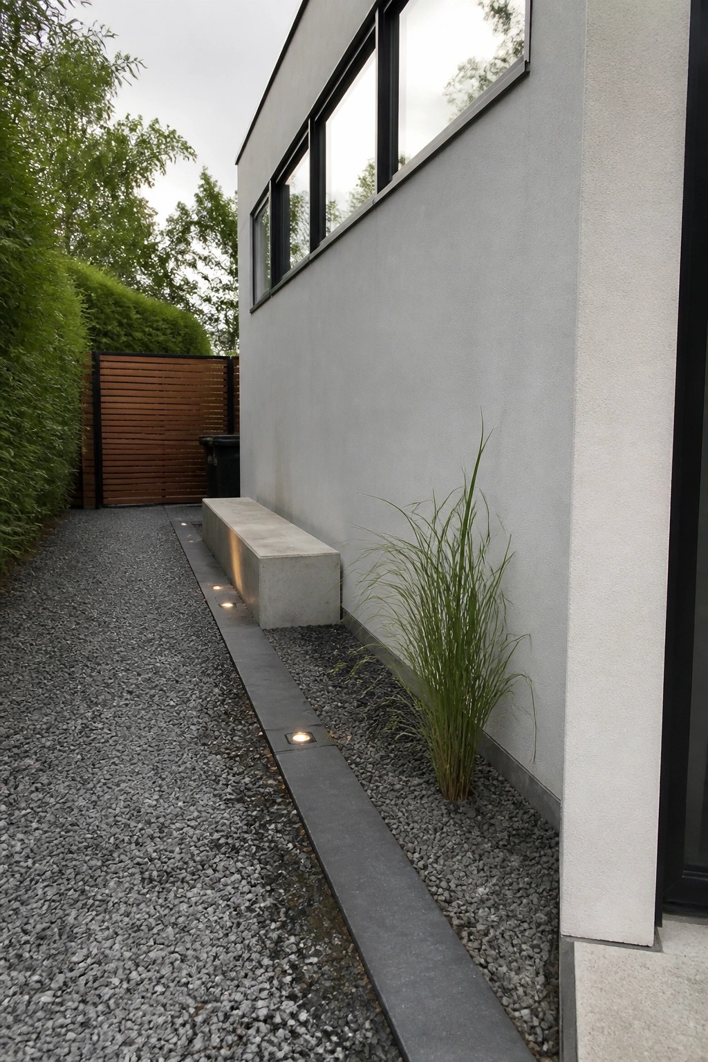 A gravel path with dark stone edging runs along the side of a gray modern house, next to a concrete bench and tall grass plantings.