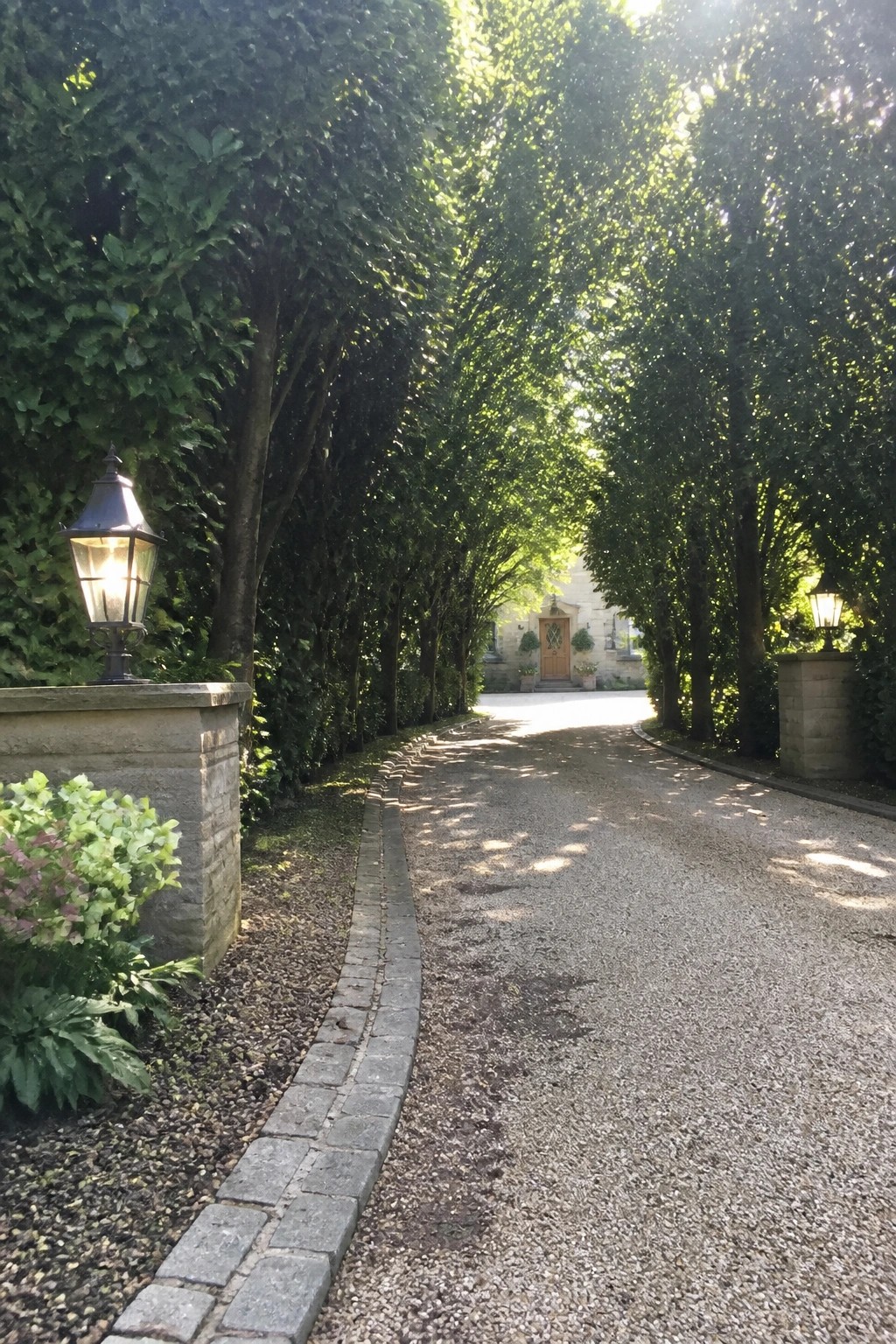 A gravel driveway with a low gray stone border curves between tall trees toward a stone house with a wooden door.