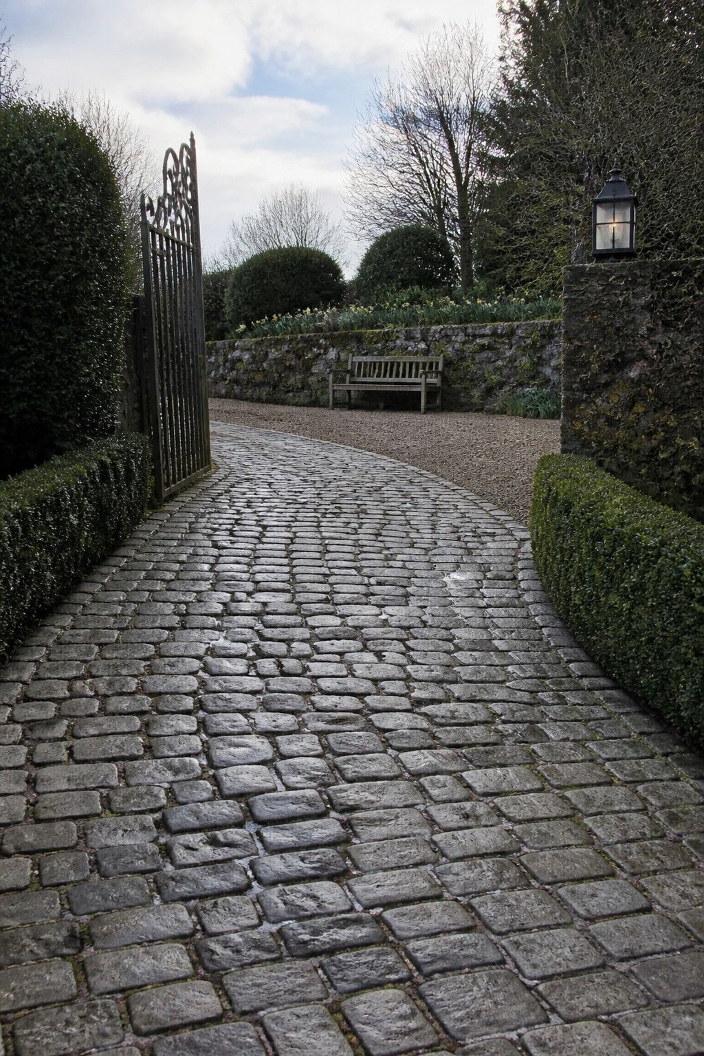 A curved cobblestone driveway passes through an open metal gate with stone pillars, hedges, and a lit lantern visible.