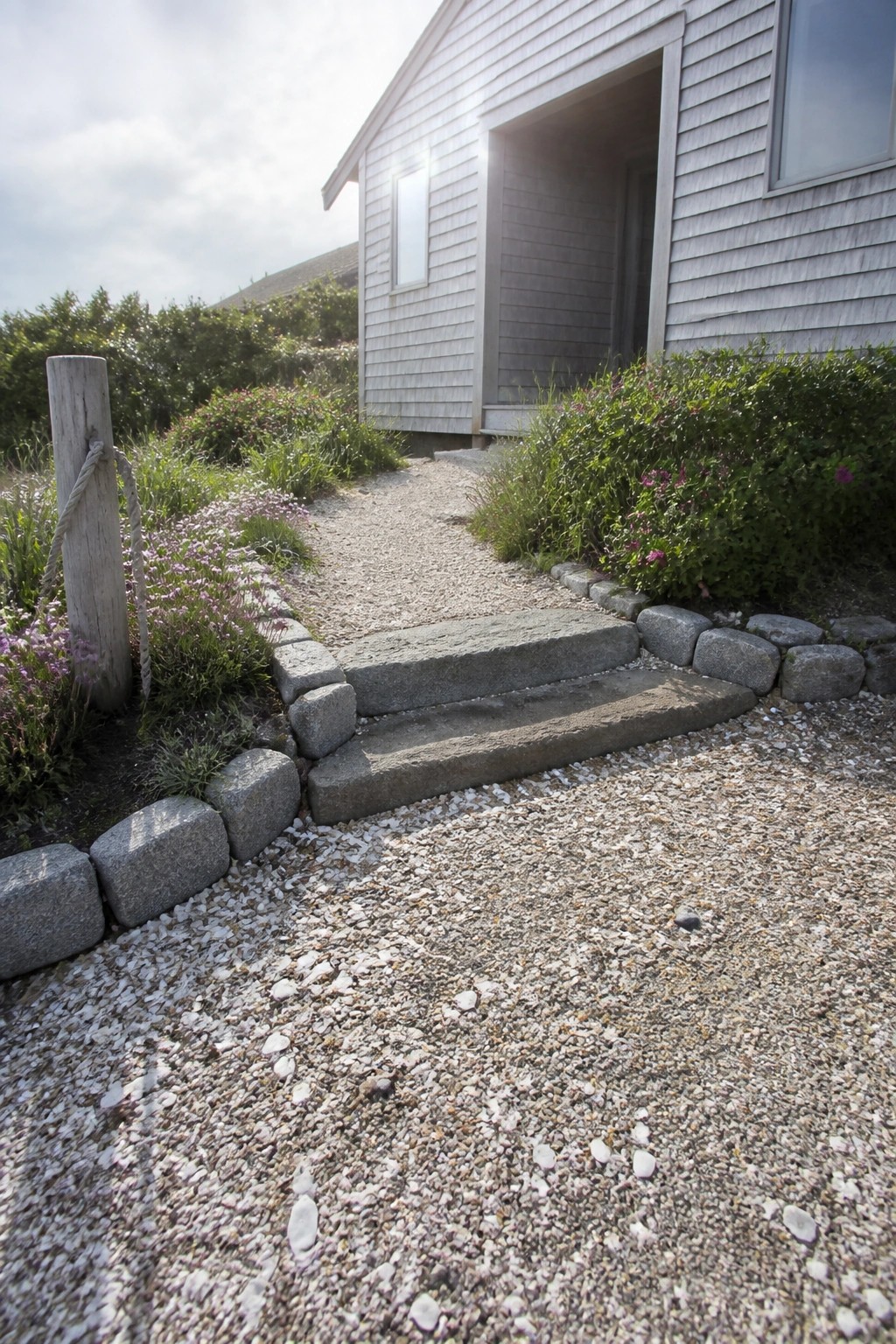 A gravel path bordered by large gray stones and purple flowering plants leads up two stone steps to the entrance of a light gray shingled house.