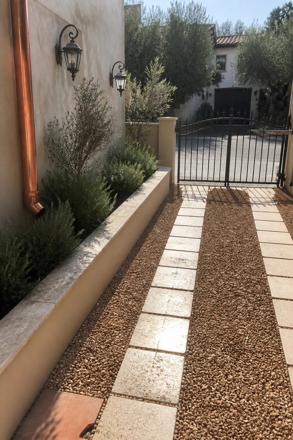 A narrow walkway made of rectangular stone pavers running through gravel, bordered on the left by a low planting bed with shrubs and a stucco wall.