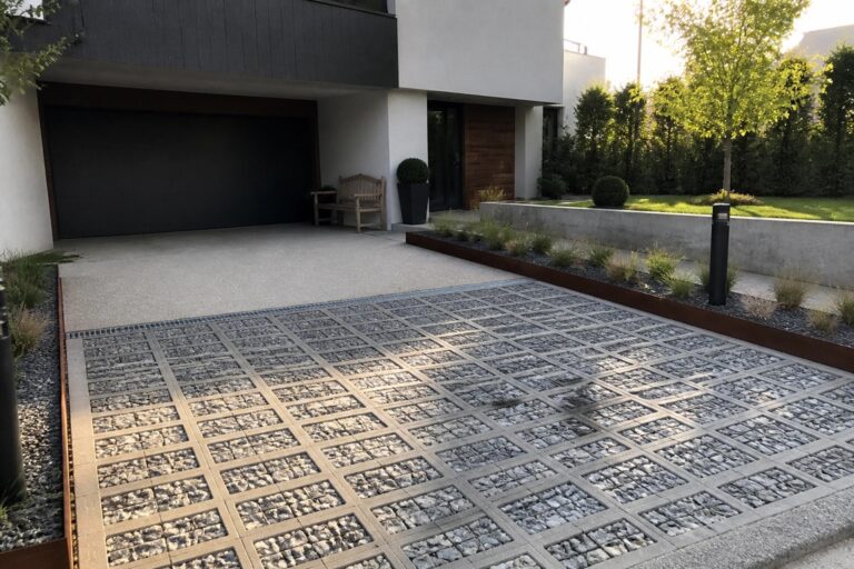 20 Durable Parking Tiles Designs Built for Heavy Use and Longevity A modern house driveway with a large section of gray concrete grid pavers filled with gravel leading to a dark garage door.