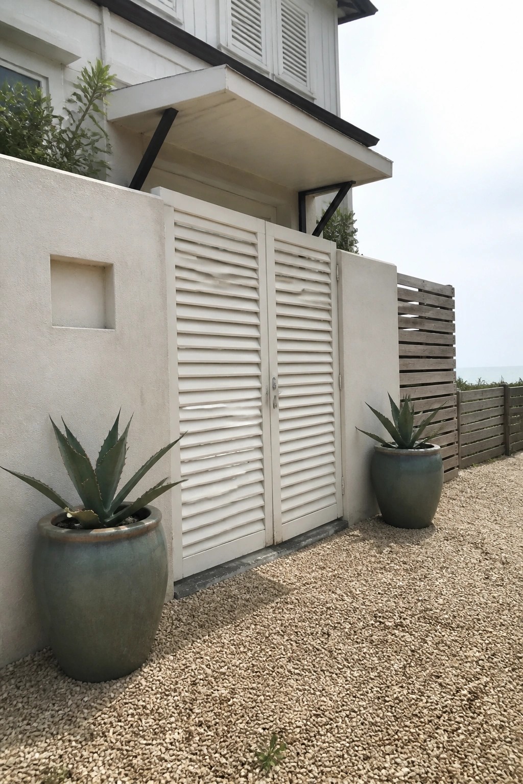 A white louvered double gate set in a beige stucco wall next to two large green ceramic pots on gravel.