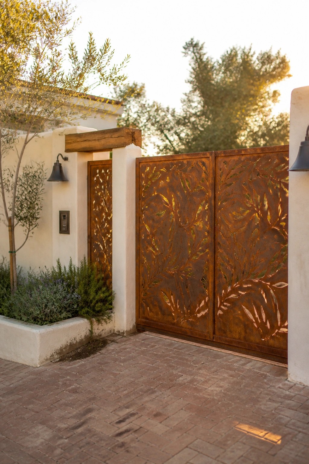 A pair of large rusted steel gates with leaf cutouts stand partially open next to white stucco walls on a brick driveway.