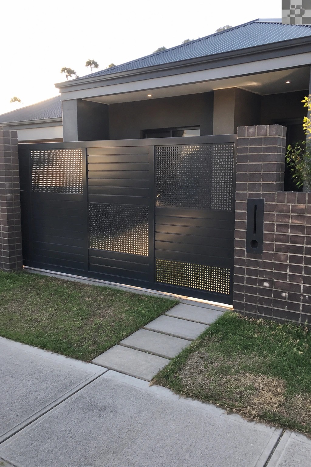 A black steel sliding gate with alternating solid horizontal panels and perforated dot sections set between brick pillars at a modern house entrance.