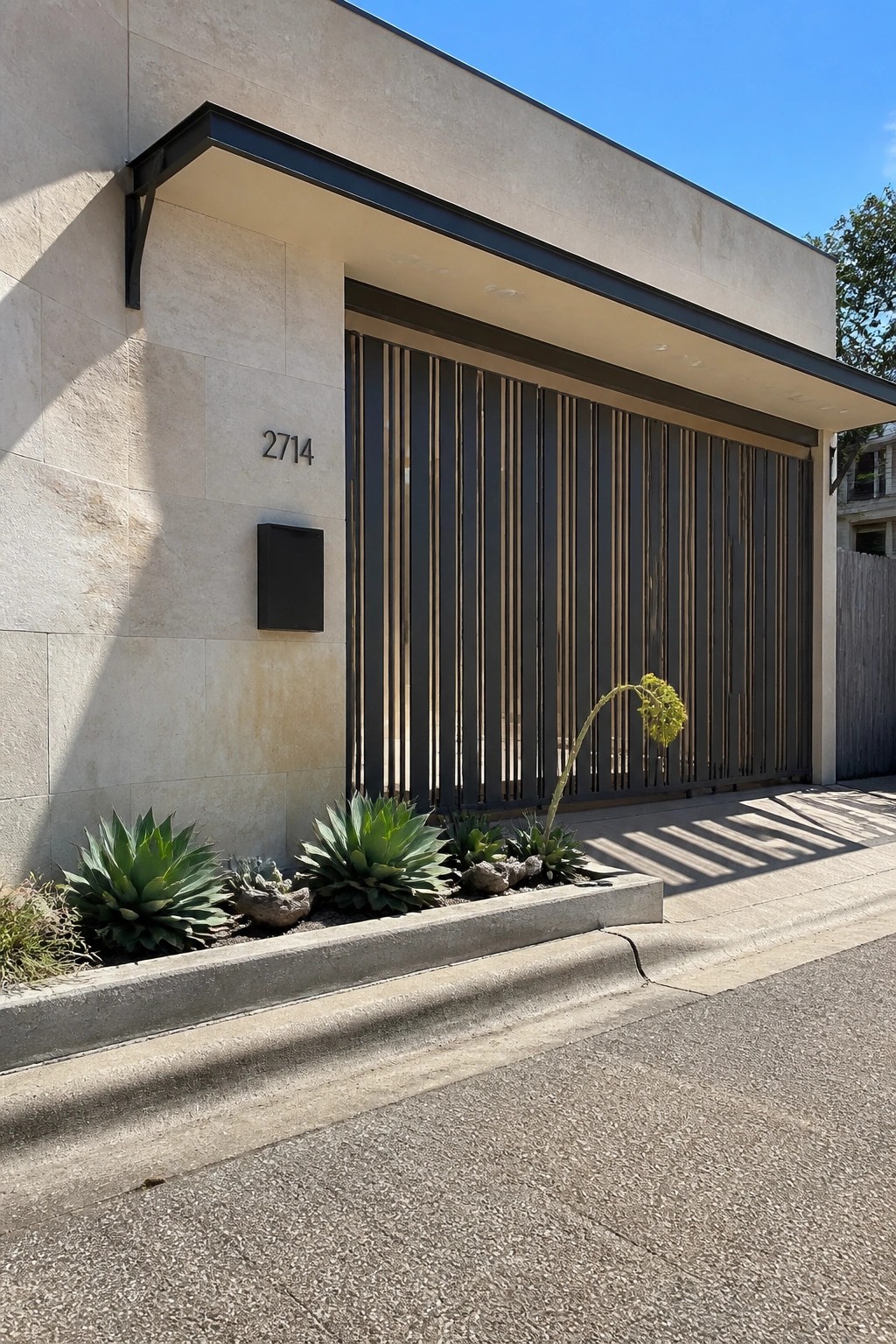 Modern house entrance featuring a dark steel gate with vertical slats beside a light stone wall with address number and agave plants.