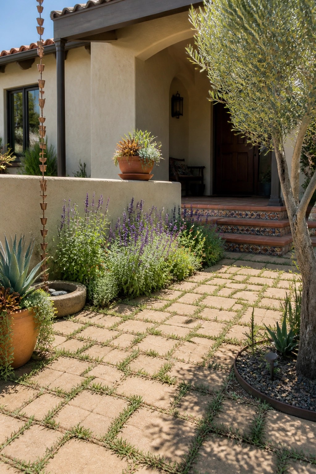 A paved walkway made of rectangular pavers with grass growing in the joints, bordered by low plants and leading toward a house entrance with tiled steps.