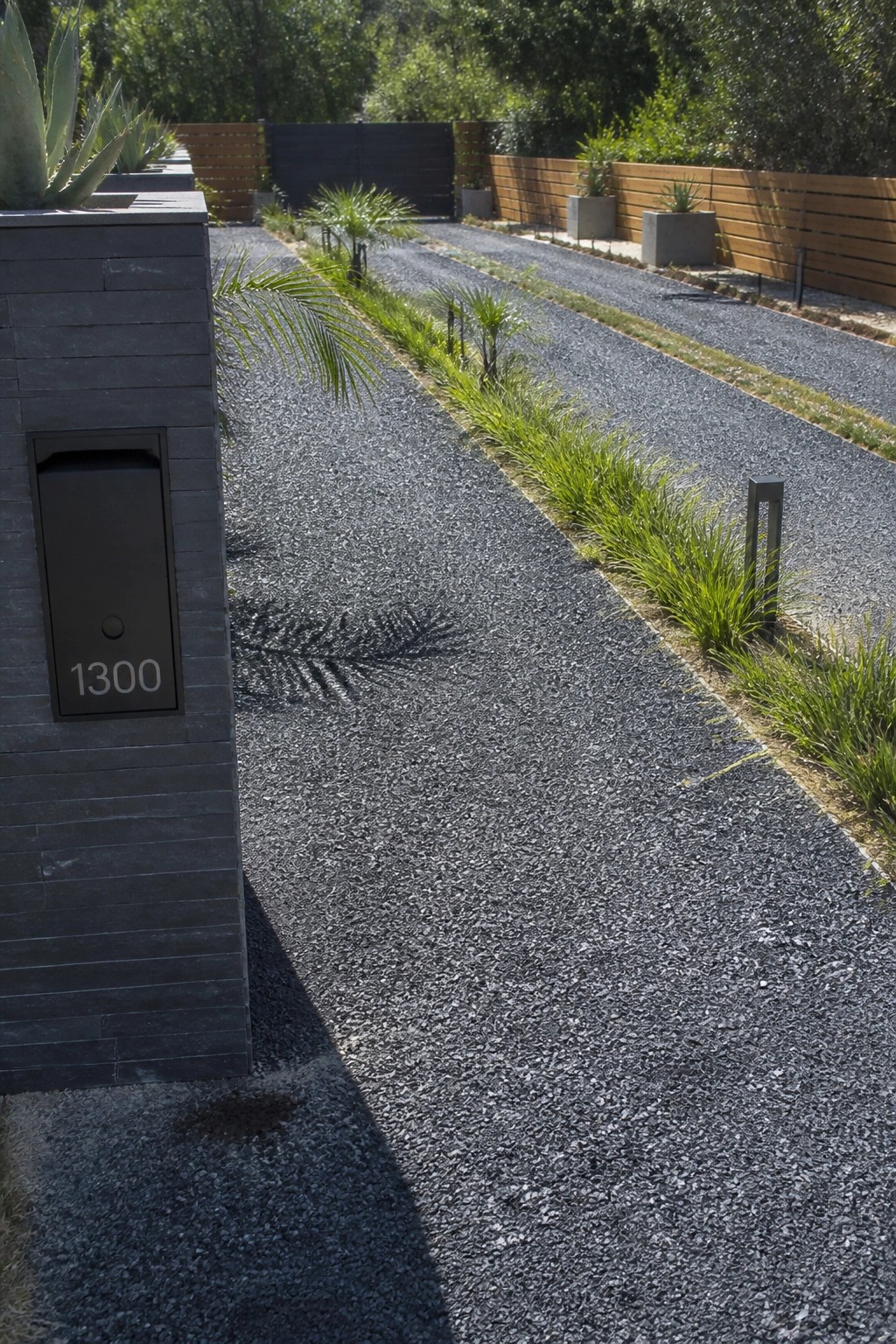 A long gravel driveway bordered by strips of green ornamental grasses, with a dark stacked stone pillar showing the address 1300 on the left side.
