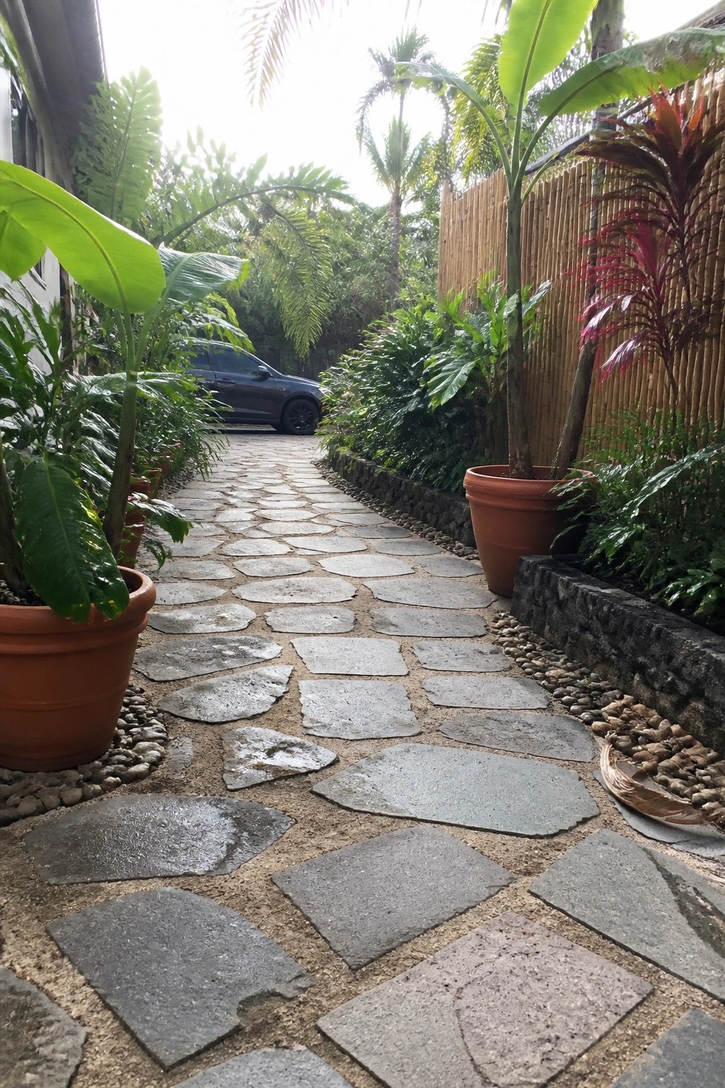 A stone paver driveway path bordered by large terracotta pots with tropical plants and gravel edging.