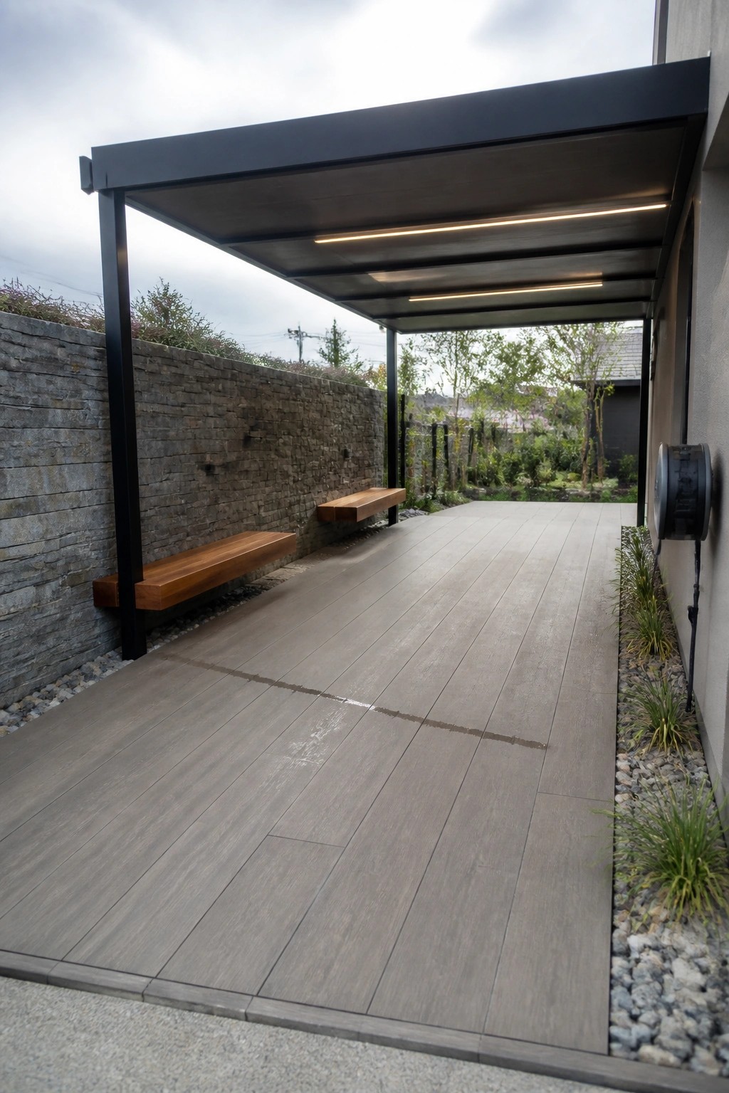 A modern covered outdoor walkway with gray wood-look plank tiles, floating wooden benches along a stacked stone wall, and gravel beds with plants on the right side.