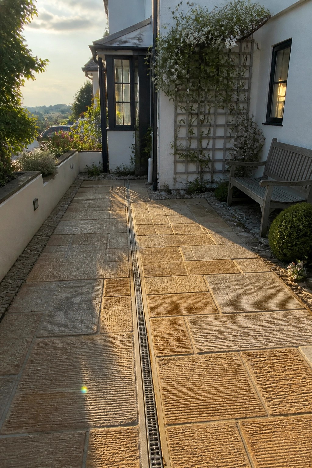 Sunlit stone path with central drain beside wooden bench and flowering trellis