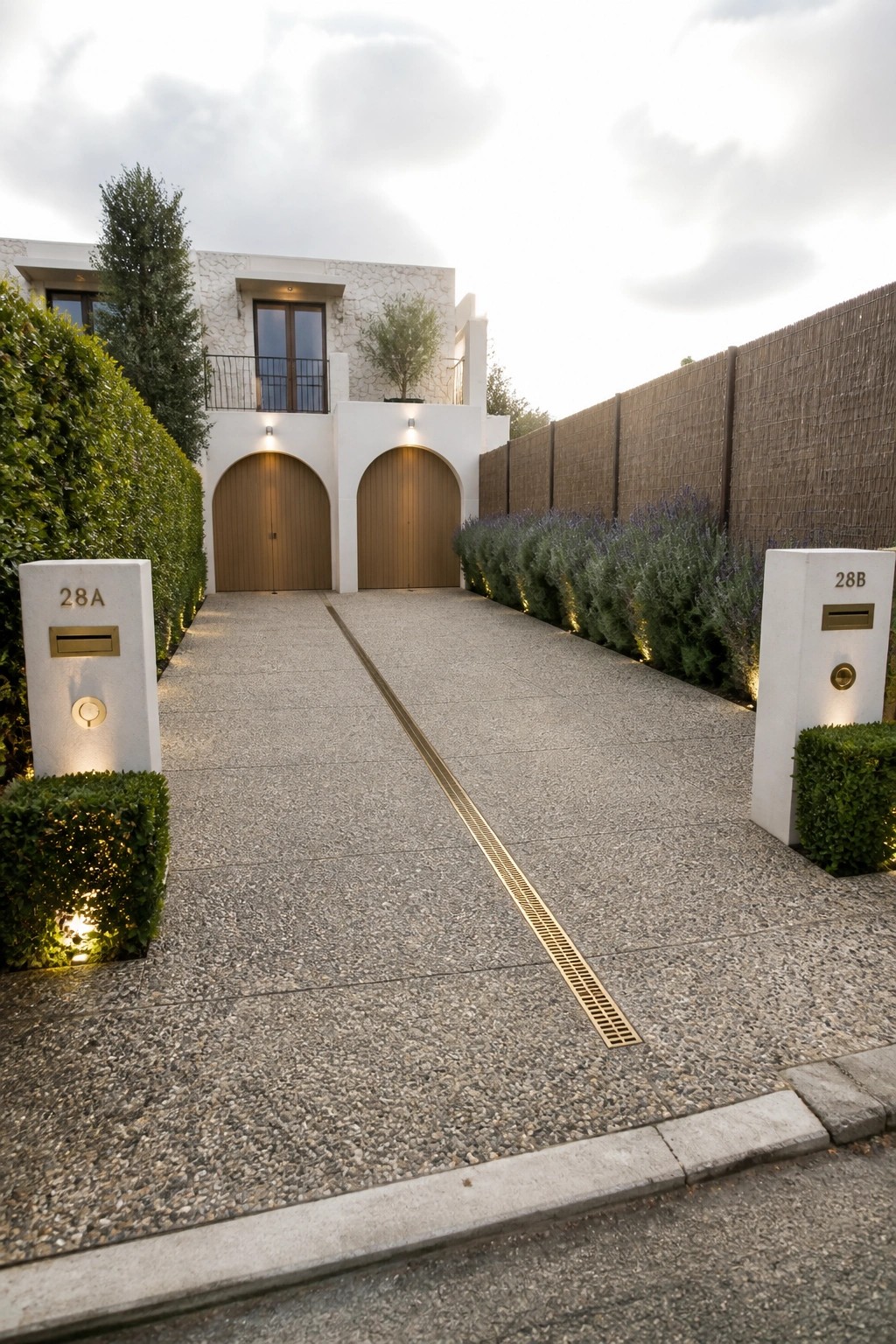 A long speckled concrete driveway with a central metal drain leads to a white house with two arched garage doors, flanked by white numbered pillars and low green landscaping.